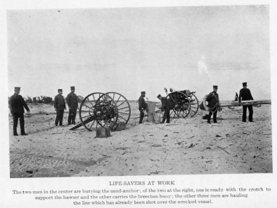 LIFE-SAVERS AT WORK
The two men in the center are burying the sand-anchor; of the two at the right, one is ready with the crotch support the hawser and the other carries the breeches-buoy; the other three men are hauling the line which has already been shot over the wrecked vessel.